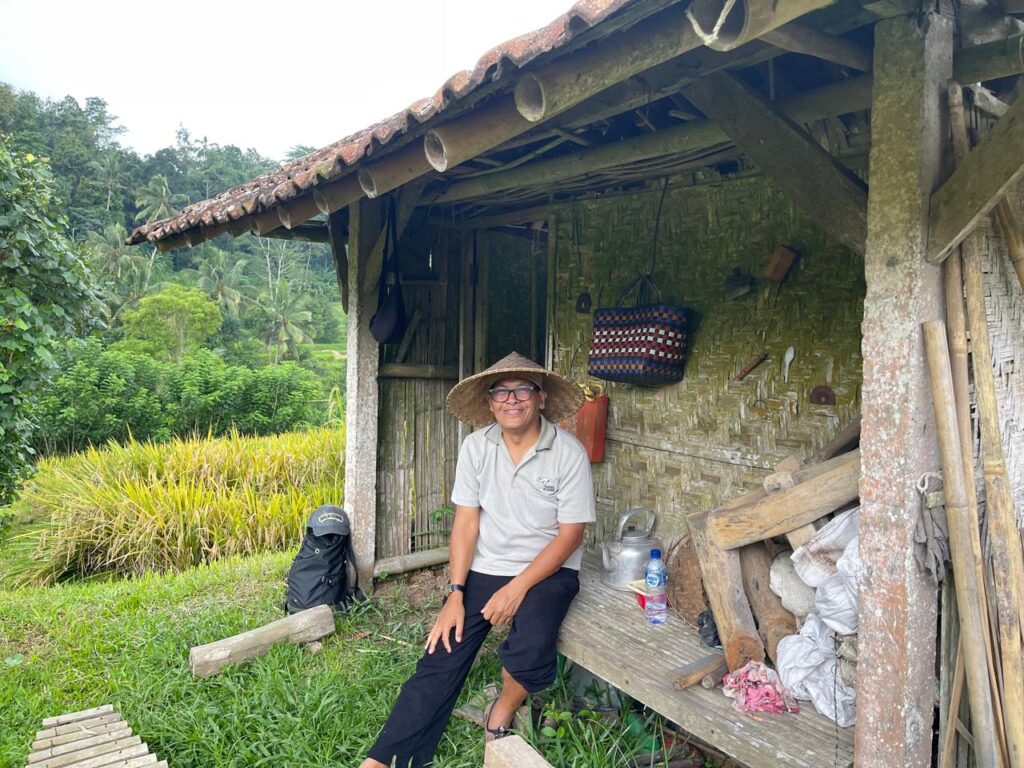 Whatsapp image 20260605 at 09 35 49 catrawarta Smiling man in a straw hat sits on a wooden bench inside a rustic shed with lush fields and rice paddies in the background